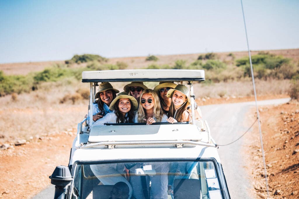 A group of six people in wide-brimmed hats pose through the pop-up roof of a white vehicle on a dusty road.