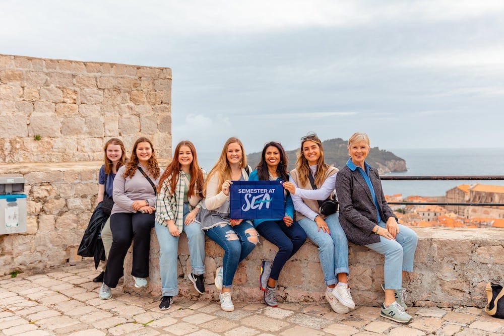 A group of seven women sit on a stone wall, smiling and holding a blue flag that reads, "Semester at Sea." Tiled rooftops and the ocean are visible in the background.