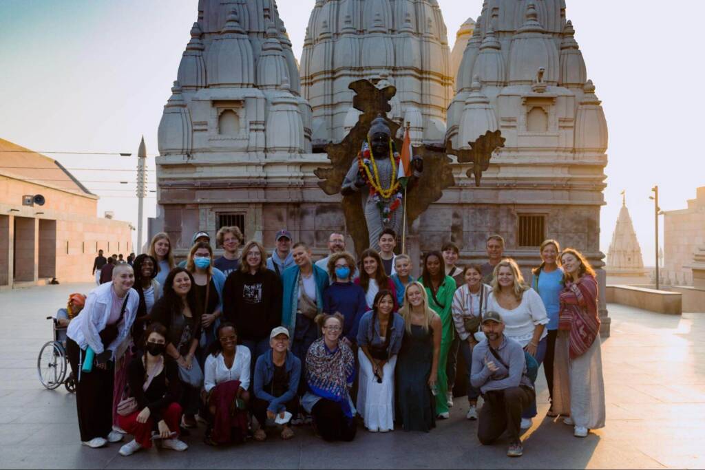 A group of about two dozen people poses in front of a life-size statue of a Hindu deity. A building with three ornately carved towers rises in the background.
