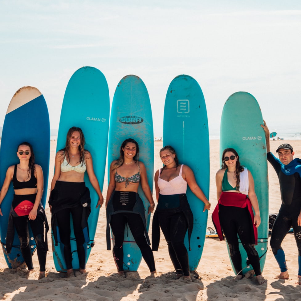 Eight smiling people stand on a beach, shoulder-to-shoulder, with five tall blue surf boards propped up in the sand behind them.