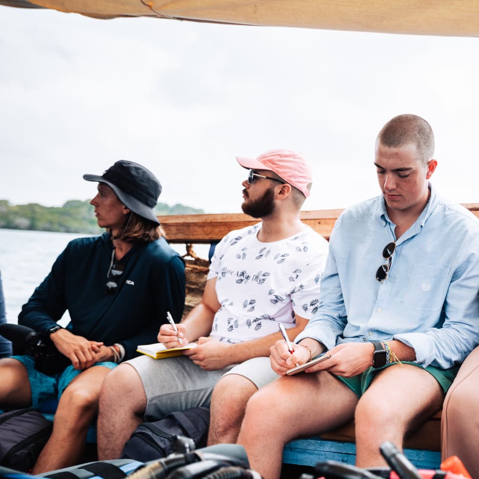 Four students sit in a boat while writing in notepads.