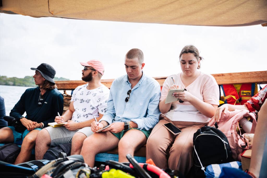 Four young people sit side-by-side on a bench against the inner wall of a wooden boat, writing in small notebooks. Water and a distant green shoreline are just visible behind them.