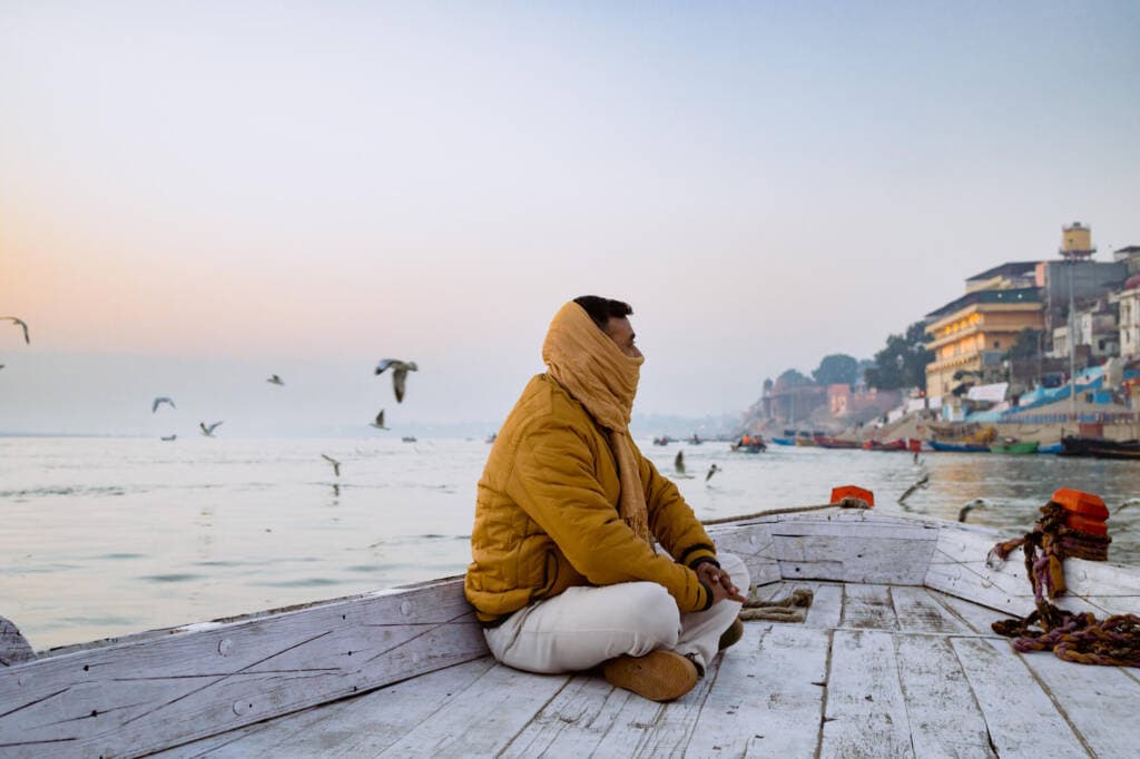 A person in a yellow jacket sits in the bow of a white, wooden boat, looking toward buildings on the shoreline, a yellow scarf wrapped around the lower half of their face. Seagulls fly over calm water in the background, which reflects pink from the evening sky.