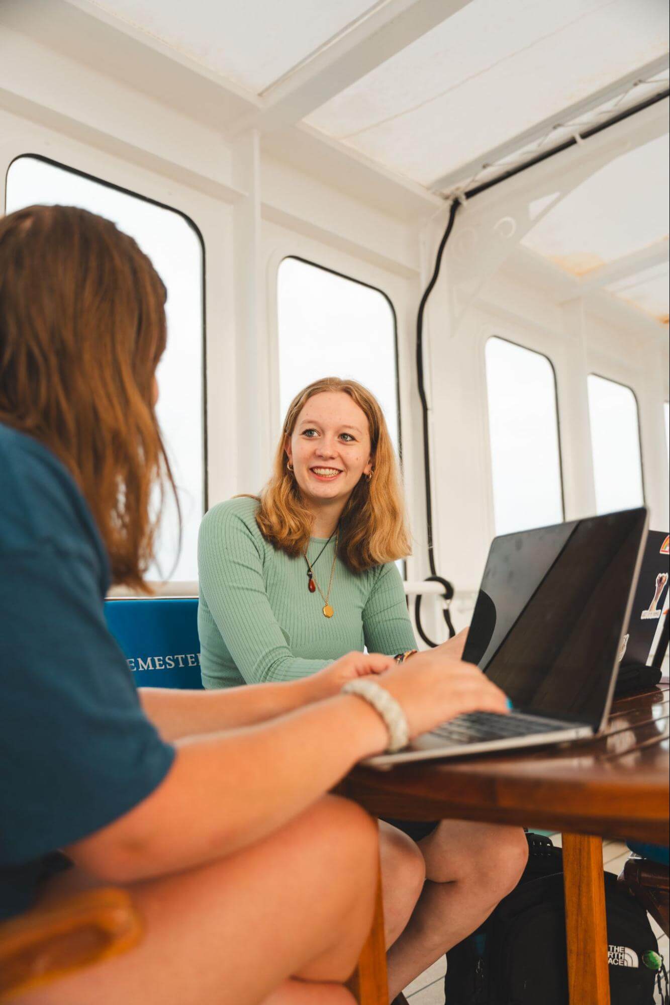 Two students sit at a table with open laptops, smiling and talking to each other. There are tall porthole windows behind them.