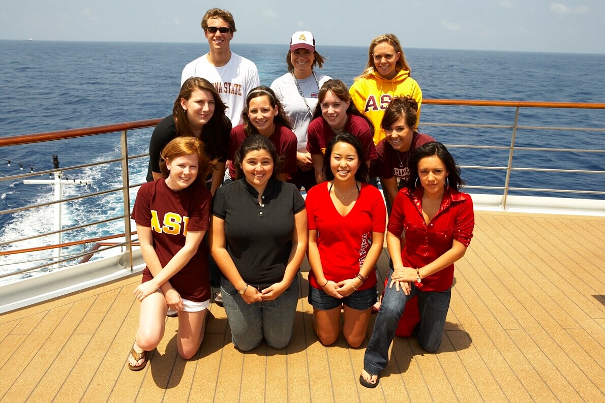 About a dozen ASU study abroad students pose in three rows, either kneeling, crouching or standing. Most are wearing a piece of ASU merchandise. They are on the sunny back deck of a ship, with the ship’s wake visible in the blue water behind them.