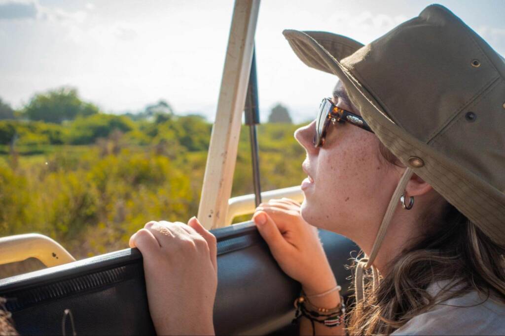 A young woman in a khaki safari hat and sunglasses grips the side of a vehicle with two hands and peers over the windowsill into the grassland beyond. 
