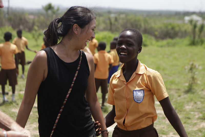 A young woman in a black tanktop turns to smile at a young girl in an orange school uniform, who reaches for the young woman’s hand and smiles at the camera. Other school children in similar uniforms play a game in the background. 